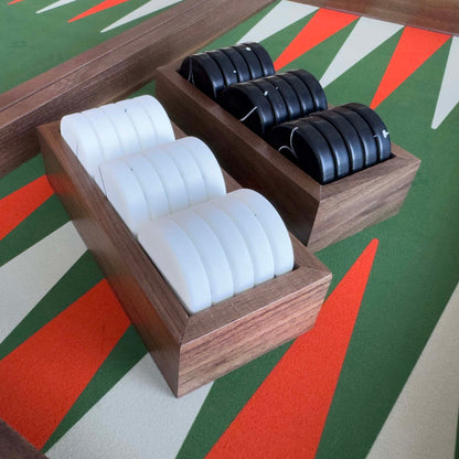Two wooden trays from the "PRIME" backgammon board holding multiple black and white cylindrical items, photographed on a colorful geometric rug.
